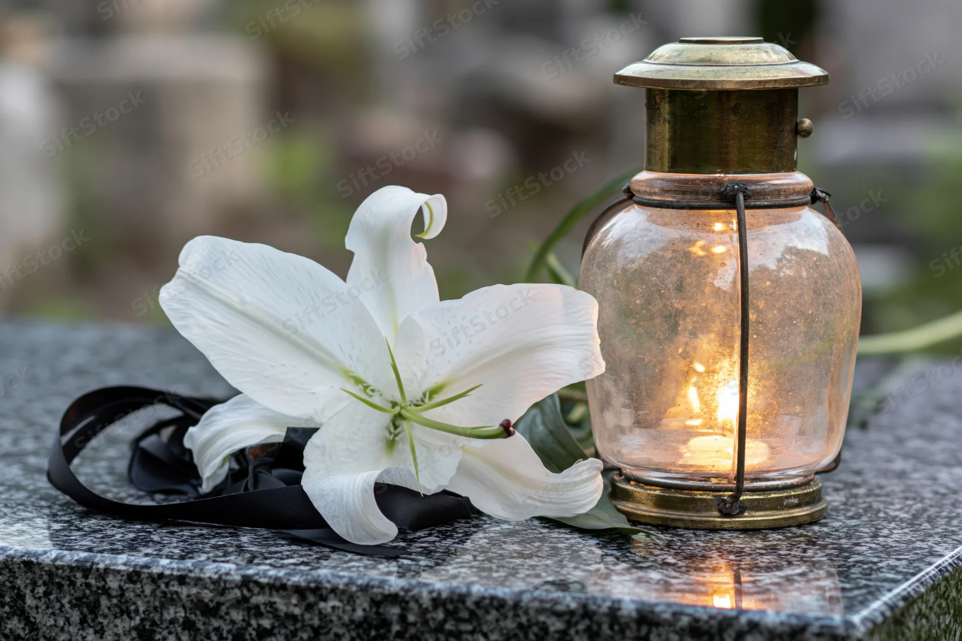 White lily with black ribbon and lit lantern on granite tombstone in cemetery