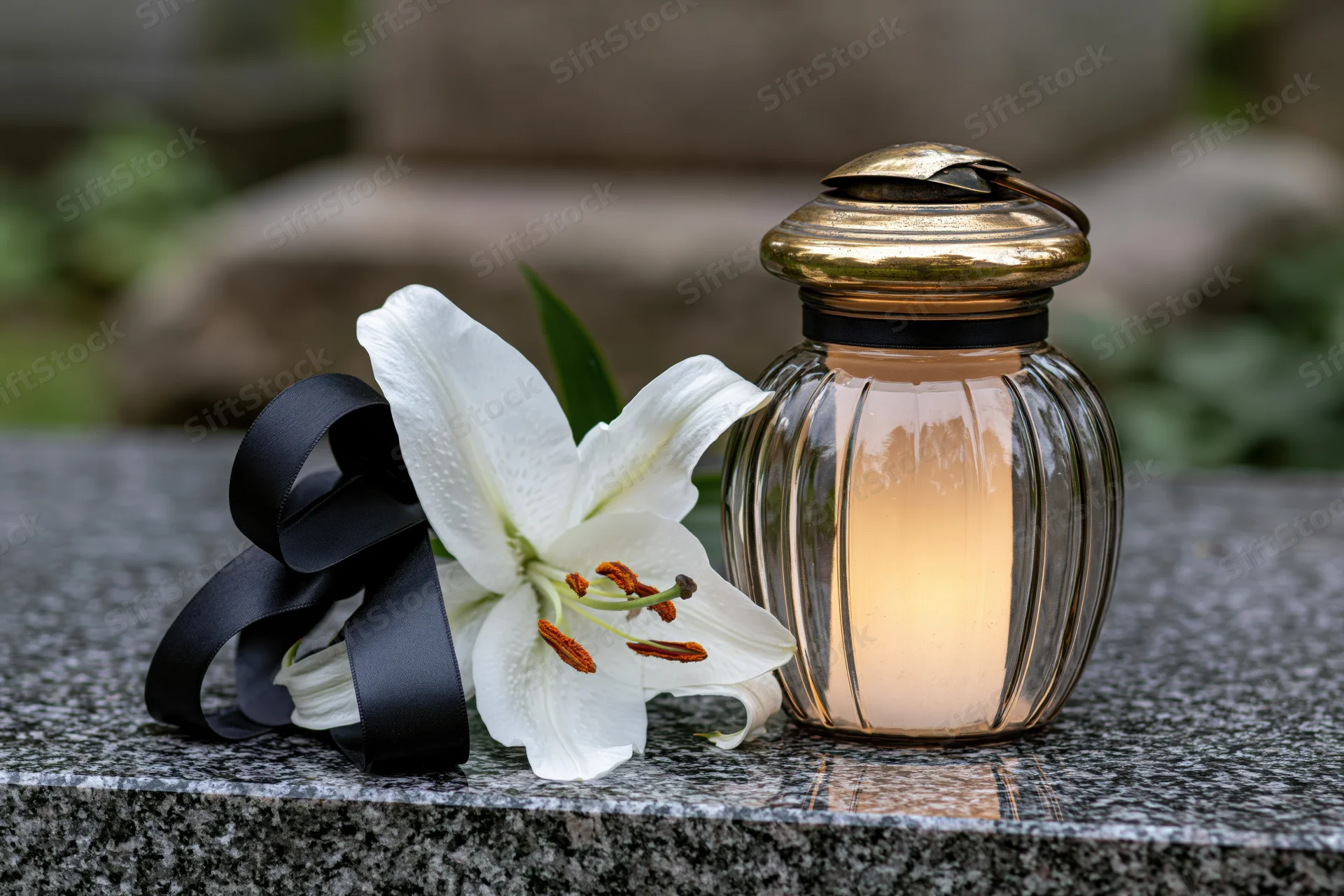 White lily with black ribbon and lit glass jar candle on granite tombstone