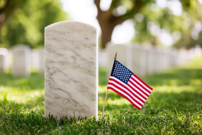 White marble military headstone in a cemetery with a small american flag planted beside it on green grass