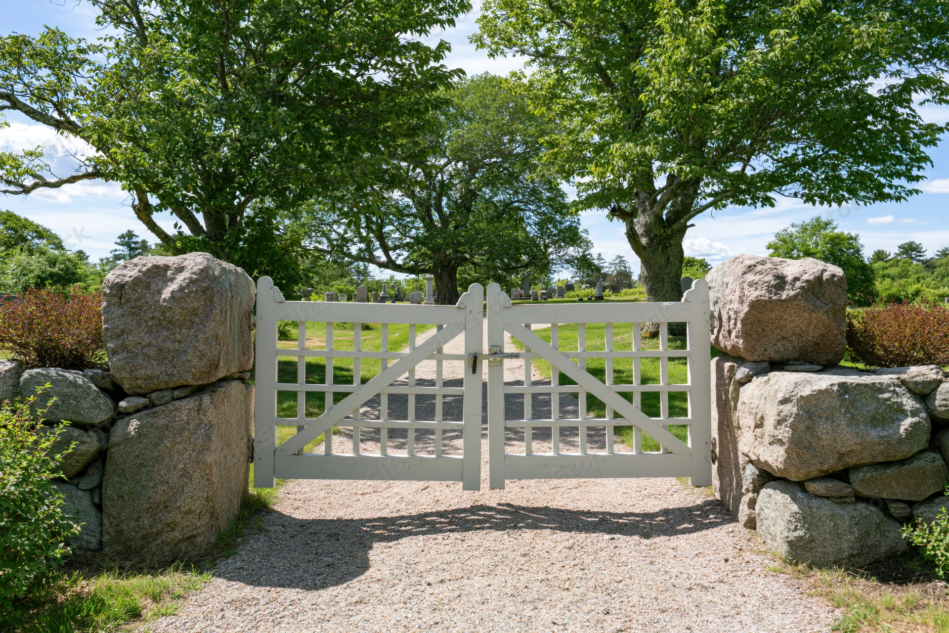White painted wooden gate with grid panels set between stone walls at cemetery entrance on sunny day