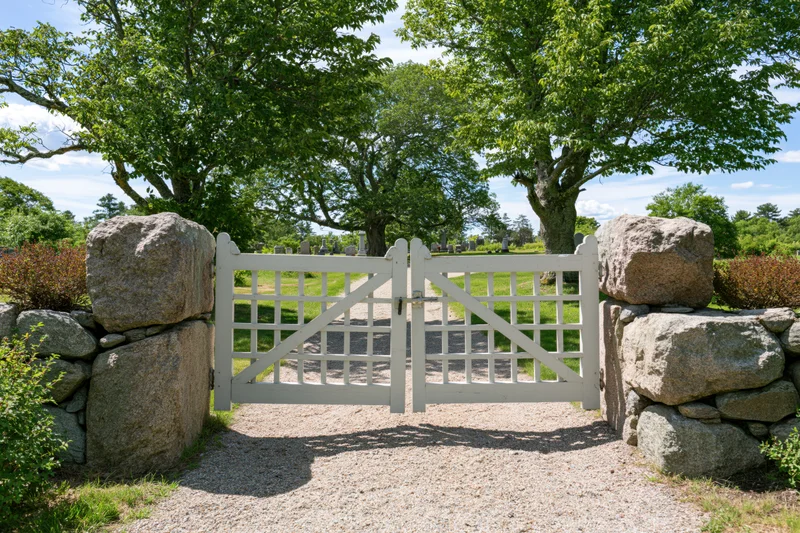 White painted wooden gate with grid panels set between stone walls at cemetery entrance on sunny day