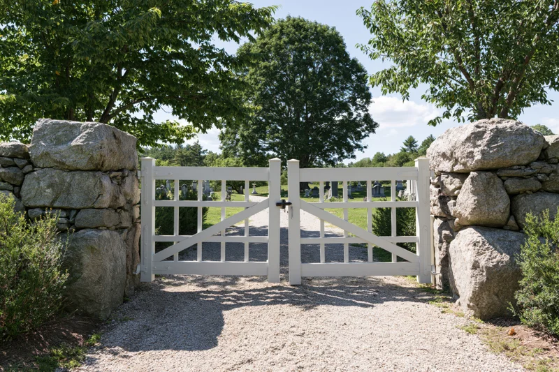White wooden gate with grid design flanked by stone walls leading to a cemetery pathway