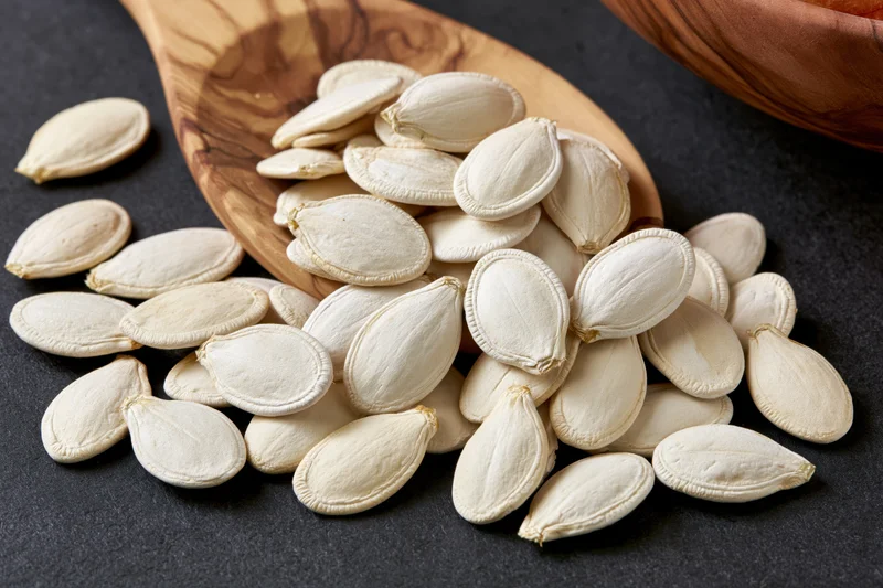 White pumpkin seeds spilling from a wooden spoon onto a dark surface