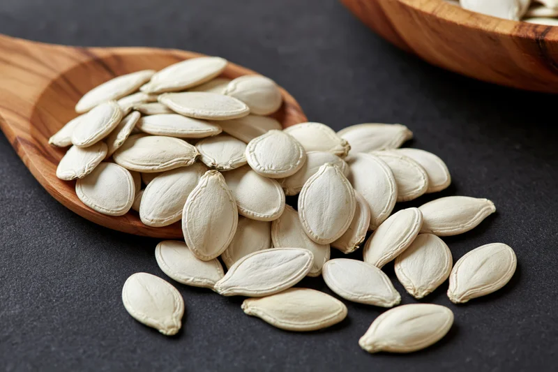 White pumpkin seeds spilling from a wooden spoon onto a dark surface