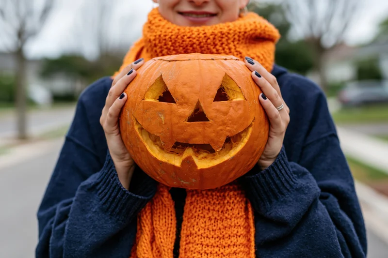 Woman holding carved jack-o-lantern in front of her face outdoors on a sidewalk