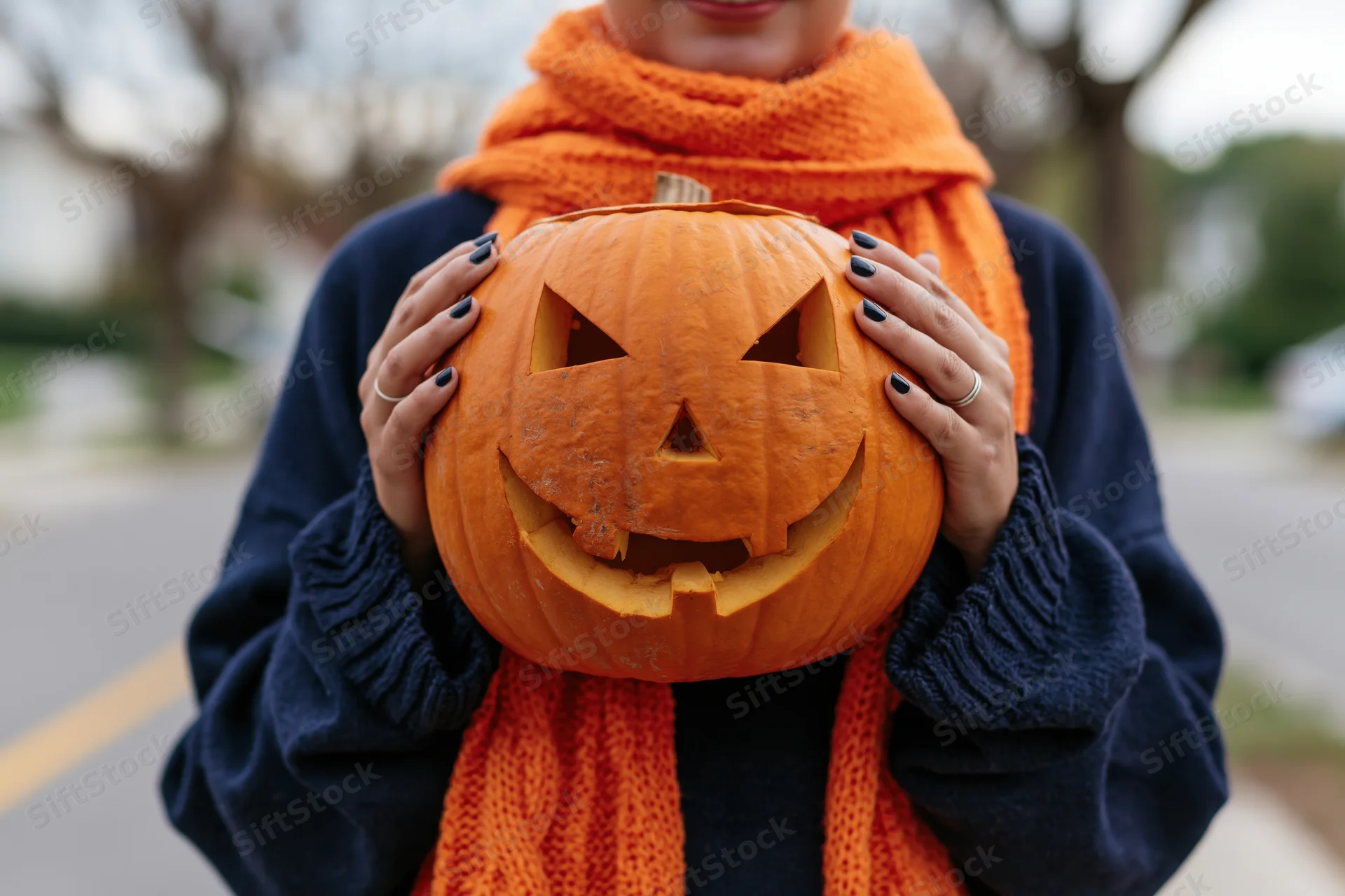 Woman holding carved jack-o-lantern in front of her chest outdoors on a street