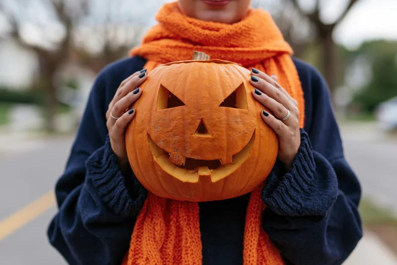 Woman holding carved jack-o-lantern in front of her chest outdoors on a street