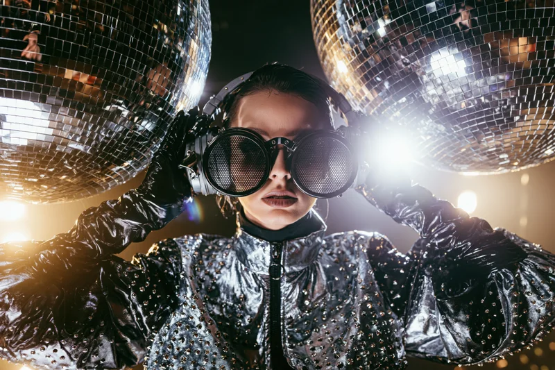 Woman in silver studded jacket and opera gloves holding goggles under disco balls at a party