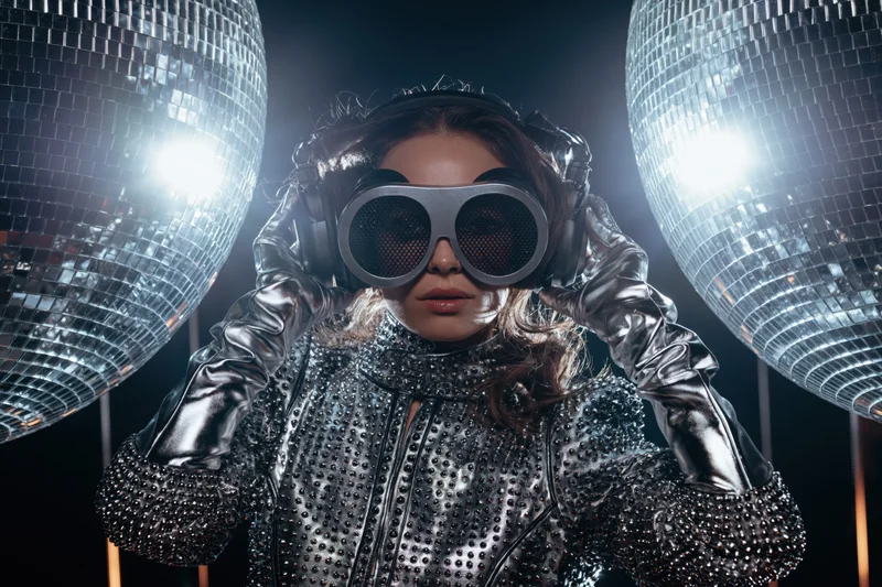 Woman in silver studded jacket and shiny opera gloves adjusting headphones under disco balls on stage