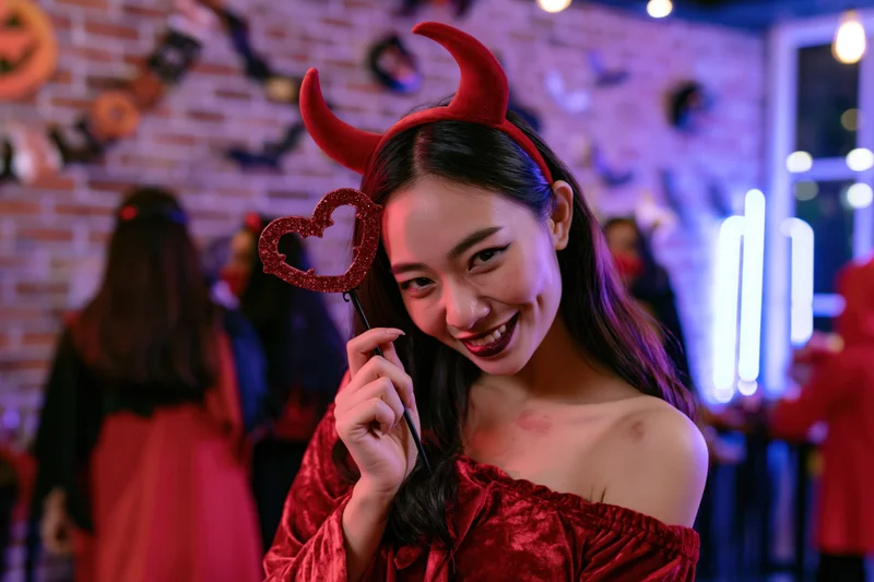 Young asian woman in red devil costume smiling with heart prop at halloween party indoors