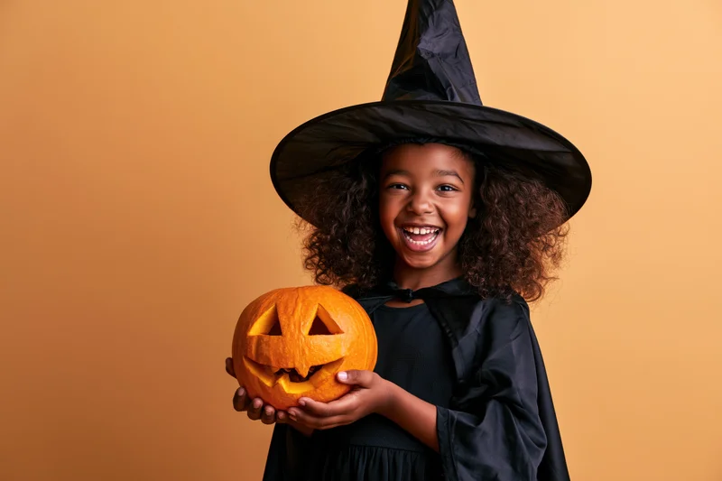 Young black girl in witch costume holding carved pumpkin, smiling against orange background