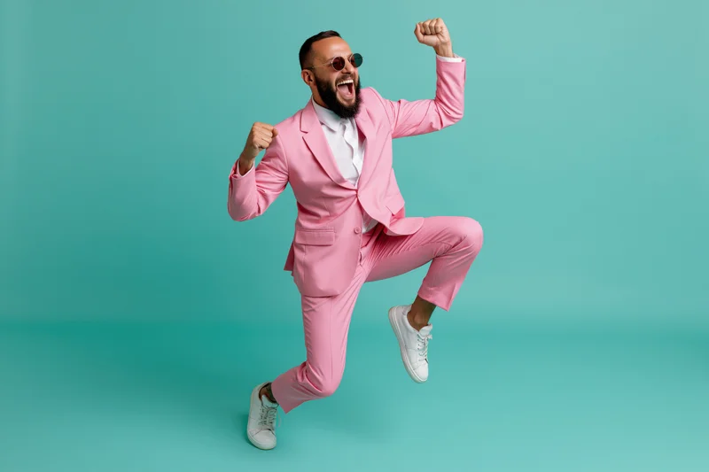 Bearded man in pink suit jumping with fists raised in celebration against turquoise background