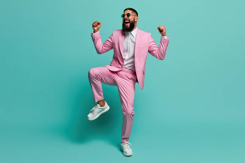 Bearded man in pink suit dancing and jumping with fists raised against turquoise background