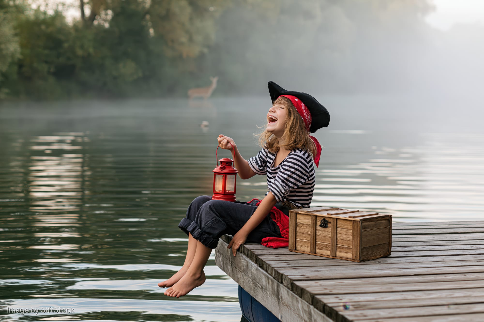 Young girl in pirate costume laughing while sitting on dock edge by misty lake