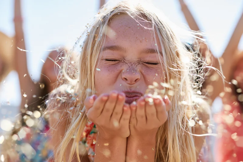 Young blonde girl blowing confetti outdoors at a festive celebration