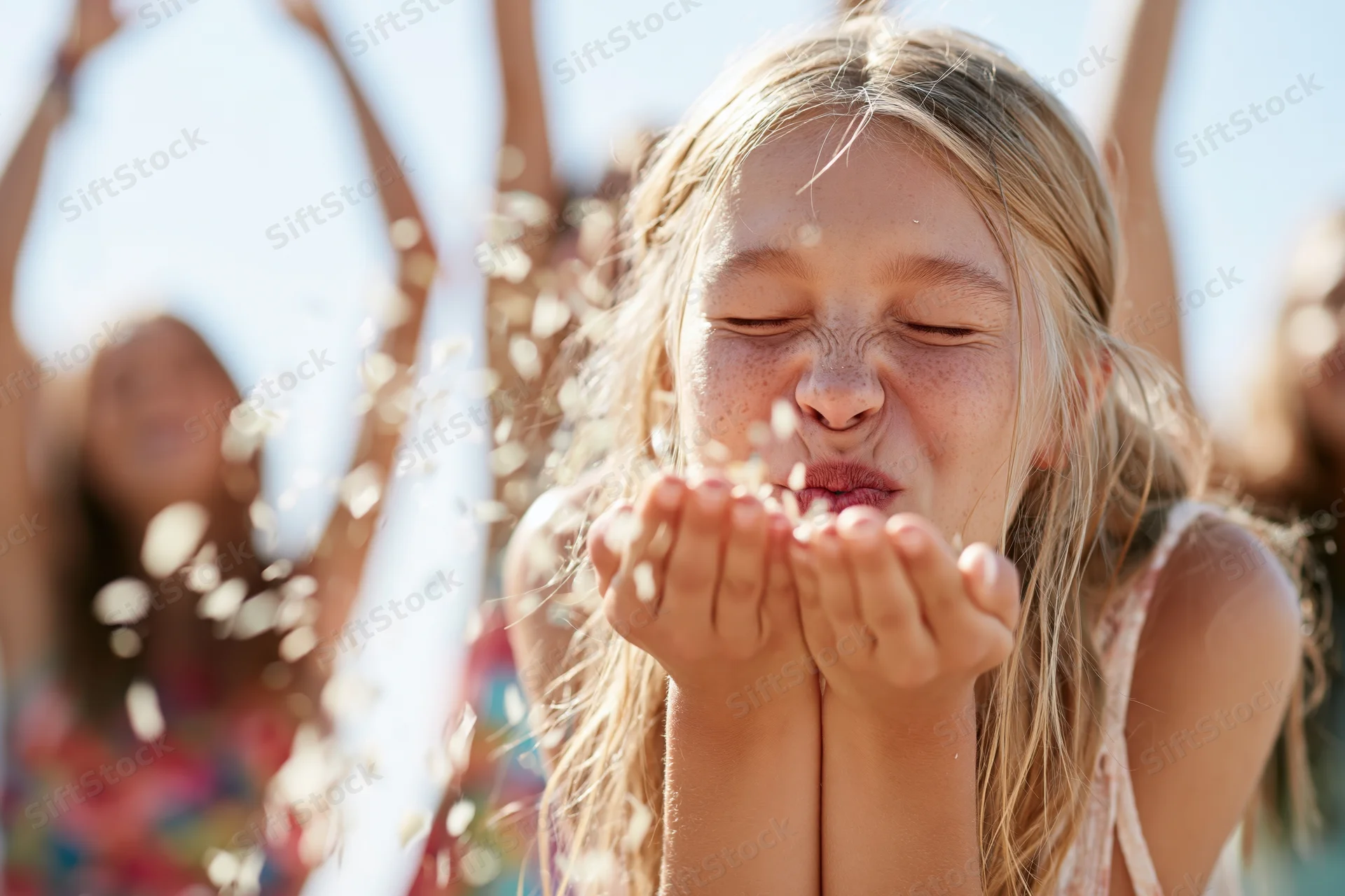 Young blonde girl blowing confetti outdoors with friends in the background