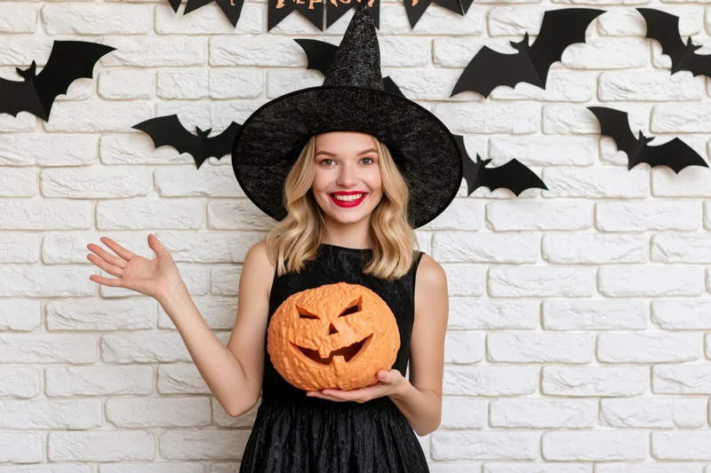 Young blonde woman in witch costume holding carved jack-o-lantern against brick wall with bat decorations