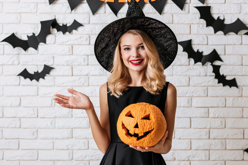 Young blonde woman in witch costume holding jack-o-lantern against decorated brick wall