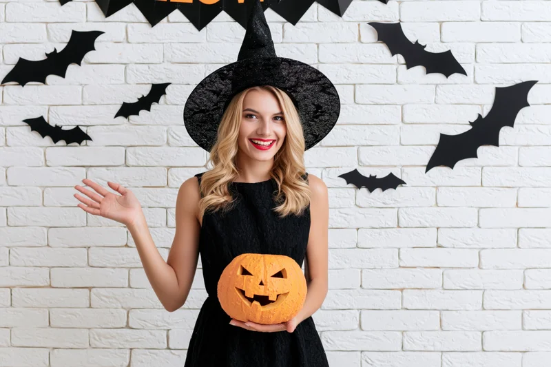 Blonde woman in witch costume holding carved jack-o-lantern against white brick wall with bat decorations