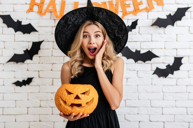 Young blonde woman in witch costume holding carved pumpkin with surprised expression in front of halloween-decorated brick wall