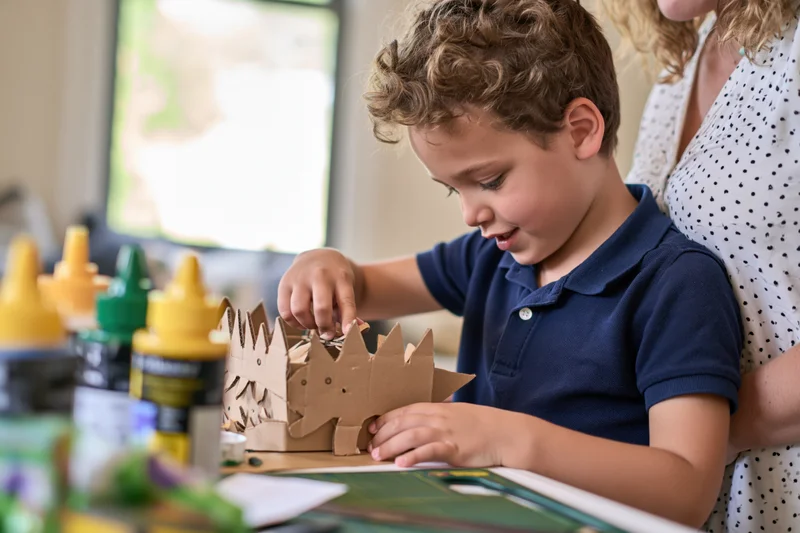 Young boy assembling a cardboard dinosaur craft at a table with glue bottles nearby while a woman supervises from behind