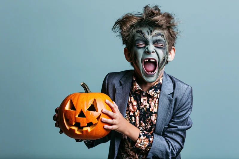 Young boy in zombie costume screaming while holding a carved jack-o-lantern against a light blue background