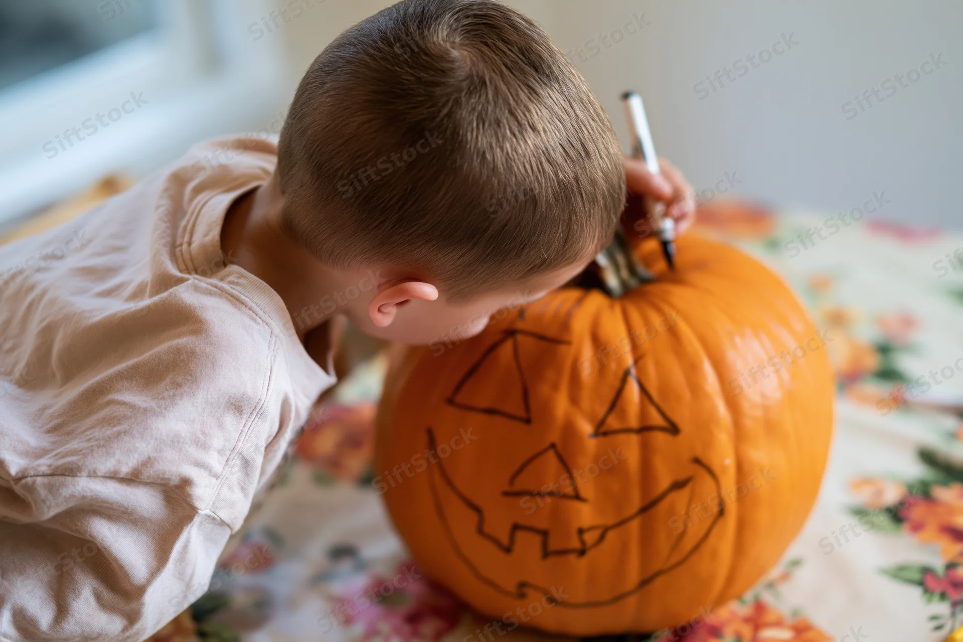 Young boy drawing a face on a pumpkin with a marker at a floral tablecloth-covered table indoors