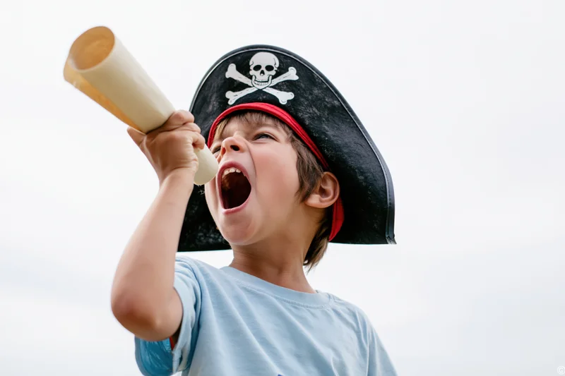 Young boy shouting into a paper tube while wearing a pirate hat outdoors