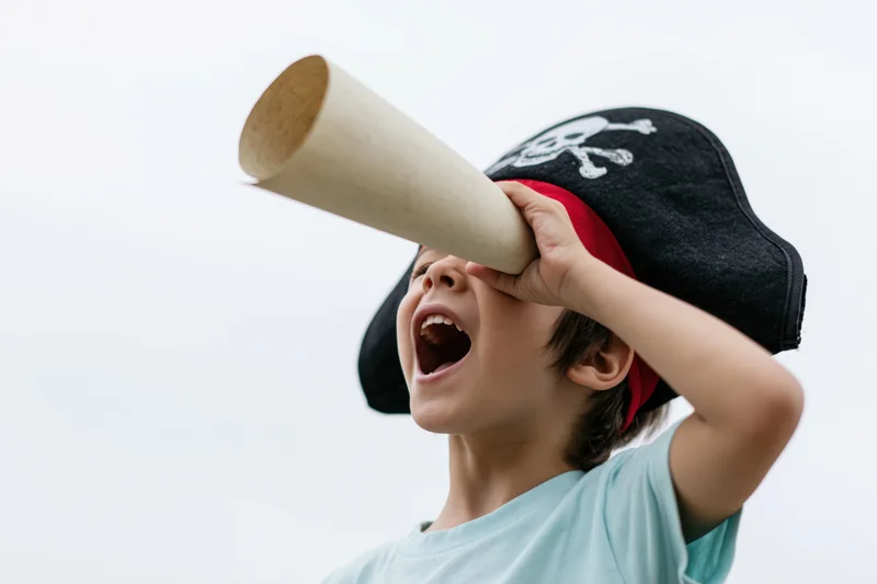 Young boy shouting into a cardboard telescope while wearing a pirate hat outdoors