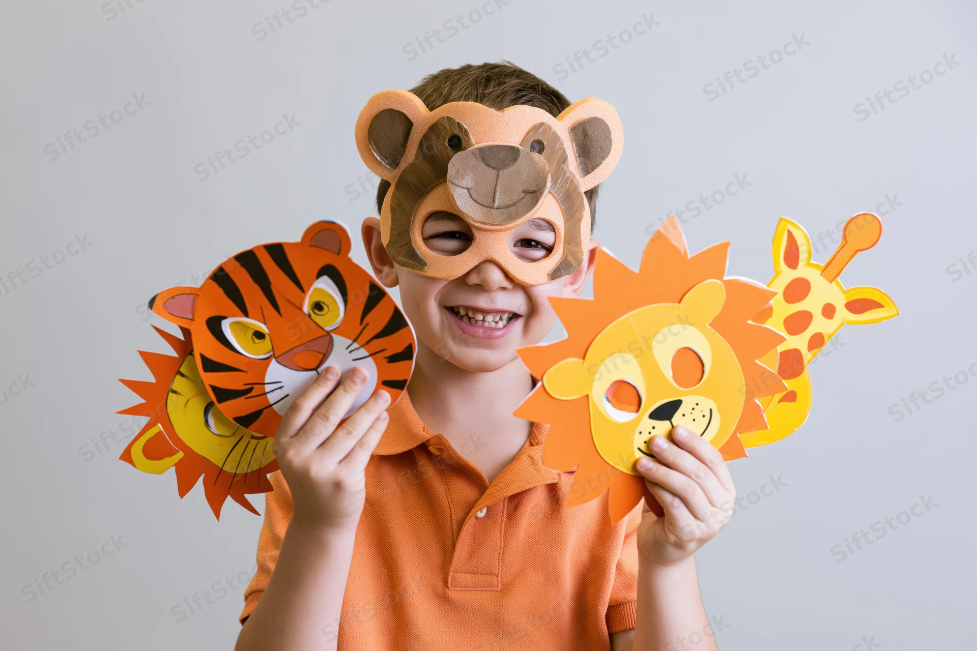 Young boy smiling while holding paper animal masks and wearing a bear mask over his eyes in a studio setting