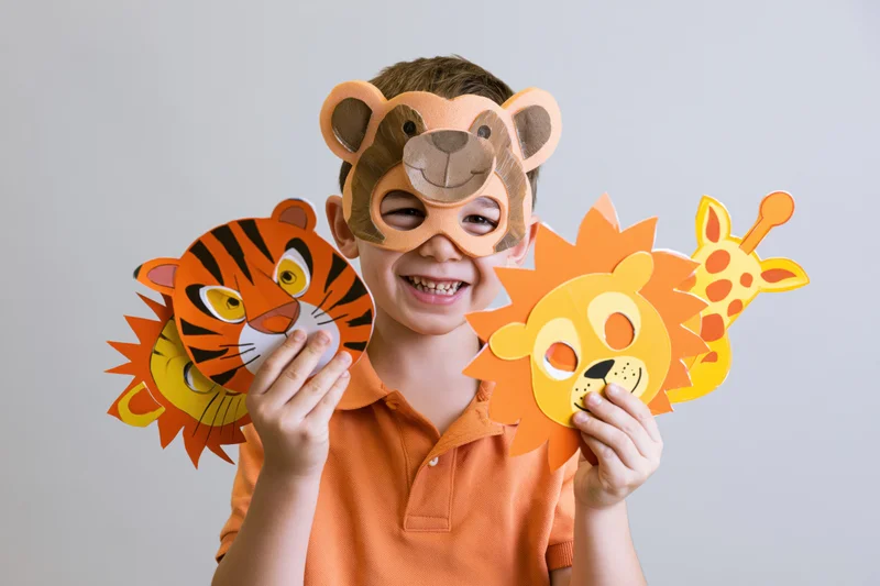Young boy smiling while holding paper animal masks and wearing a bear mask over his eyes in a studio setting
