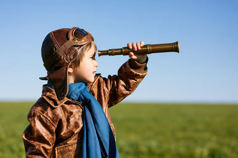 Young child in vintage aviator costume holding telescope outdoors in a green field under blue sky