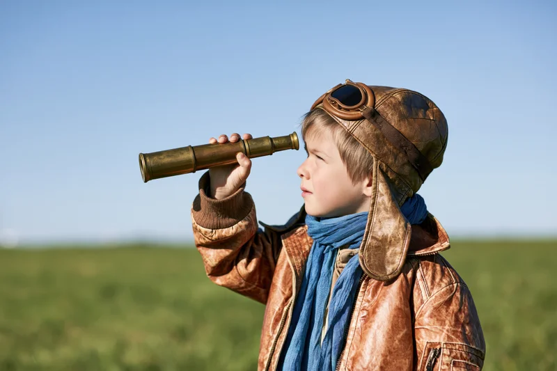 Young child in vintage aviator costume holding brass telescope outdoors in a grassy field under blue sky