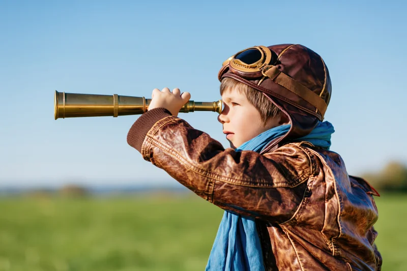 Young boy in vintage aviator costume looking through brass telescope in a green field under blue sky