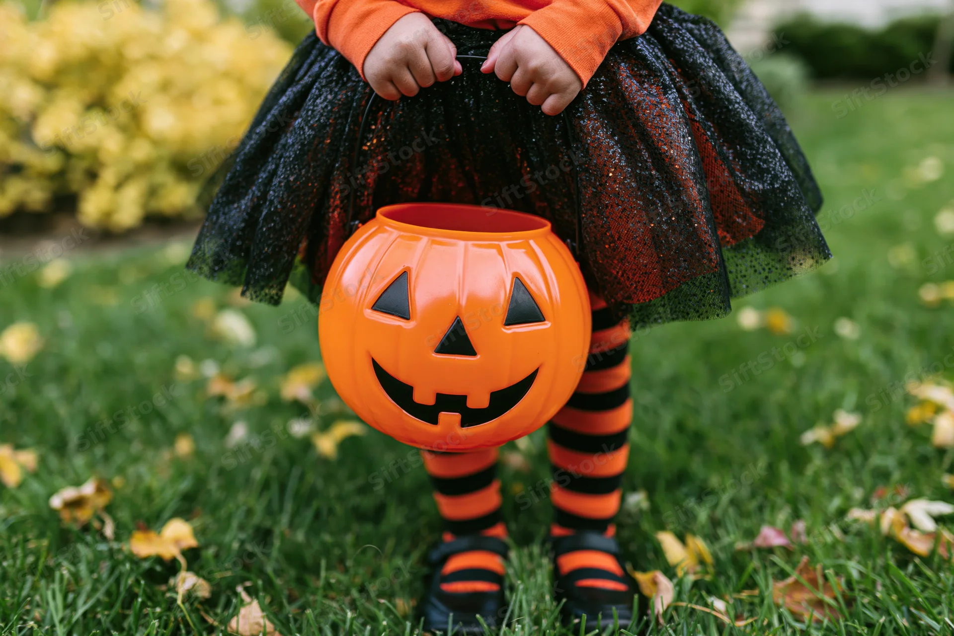Young girl in halloween costume holding orange jack-o-lantern candy bucket on grassy yard