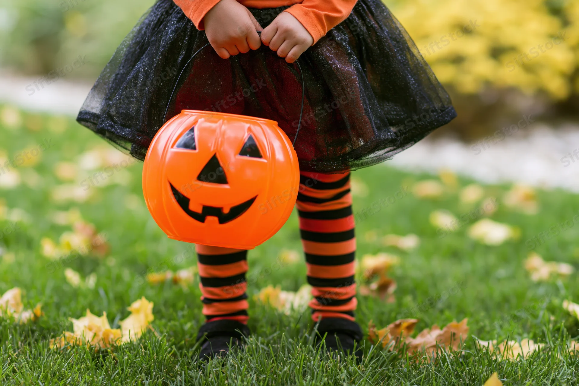 Child holding orange jack-o-lantern candy bucket on grass with autumn leaves