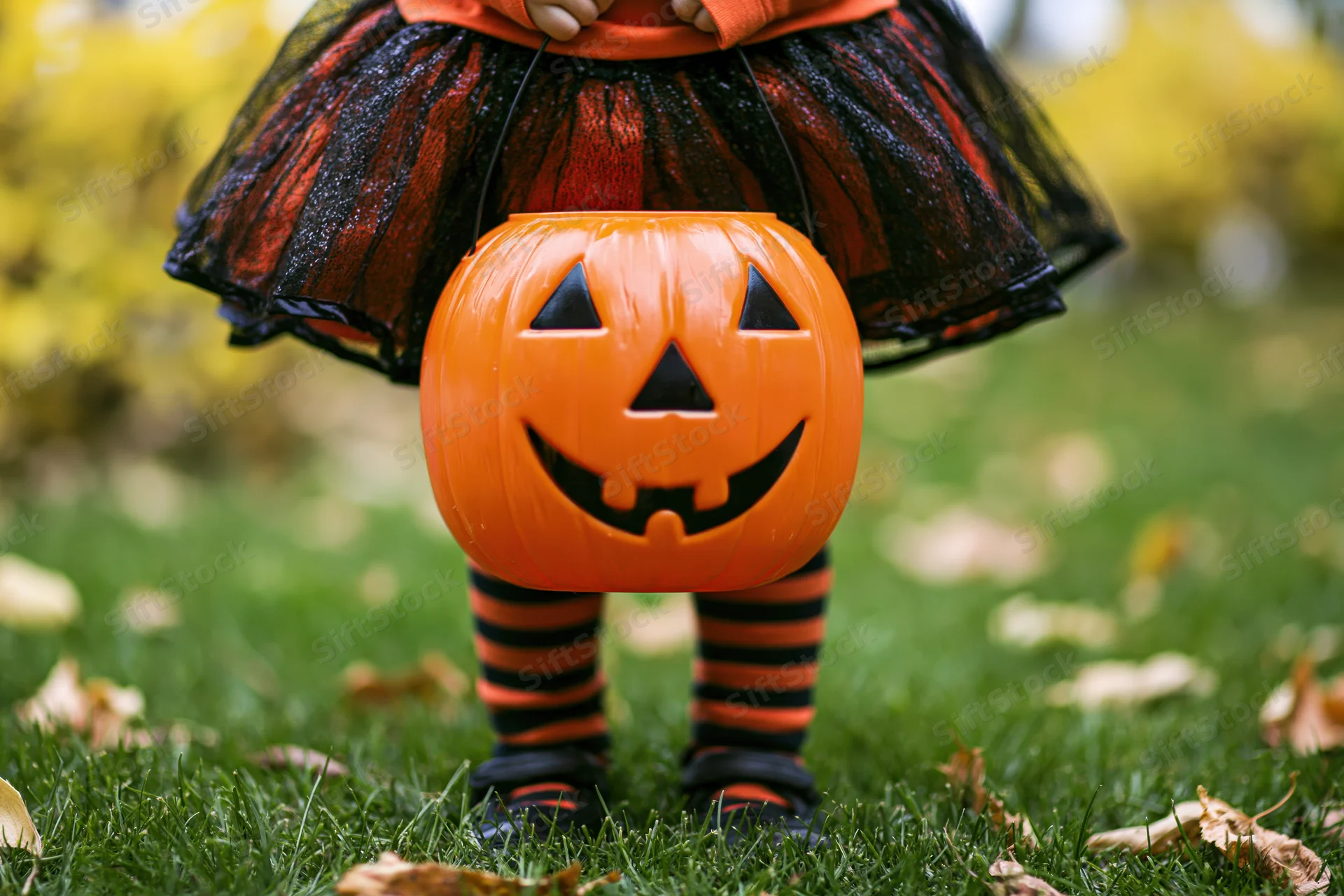 Young child in halloween costume holding orange jack-o-lantern candy bucket on grassy lawn