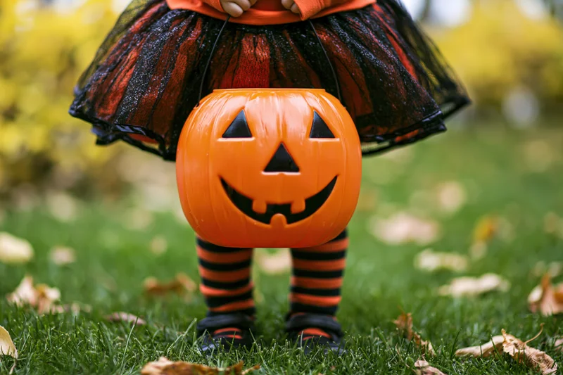 Young child in halloween costume holding orange jack-o-lantern candy bucket on grassy lawn