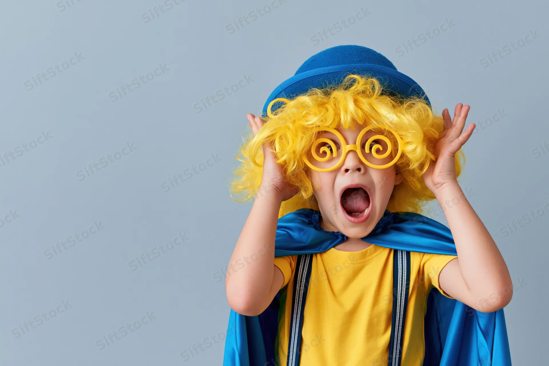 Child in clown costume with yellow wig and spiral glasses making exaggerated surprise expression against blue background