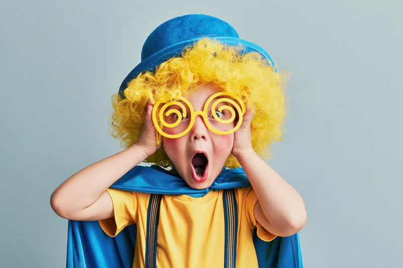 Child in clown costume with yellow wig and spiral glasses making exaggerated surprise expression