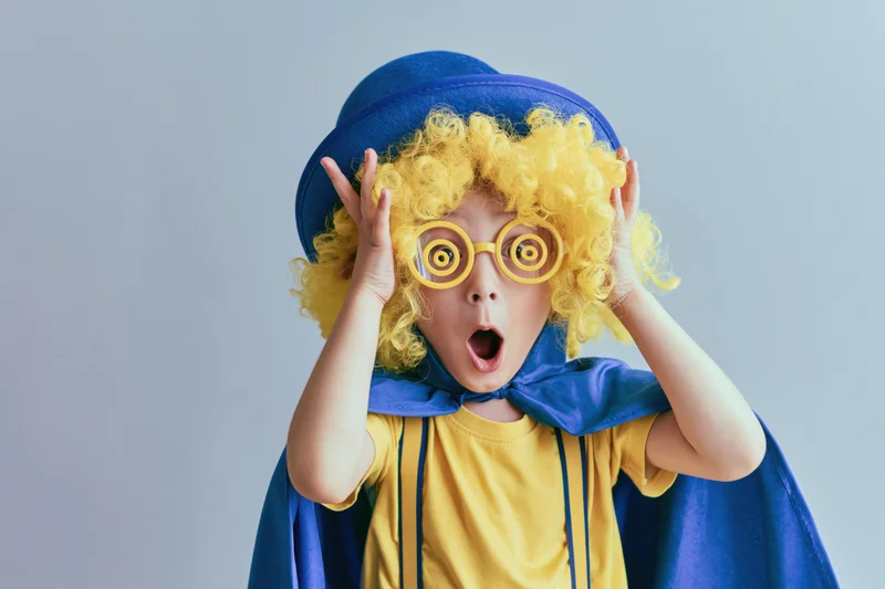 Child in clown costume with yellow wig and blue hat making exaggerated surprise expression in studio