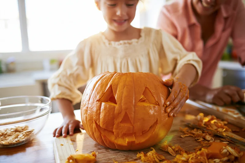 Young girl carving a jack-o-lantern with an adult in a kitchen