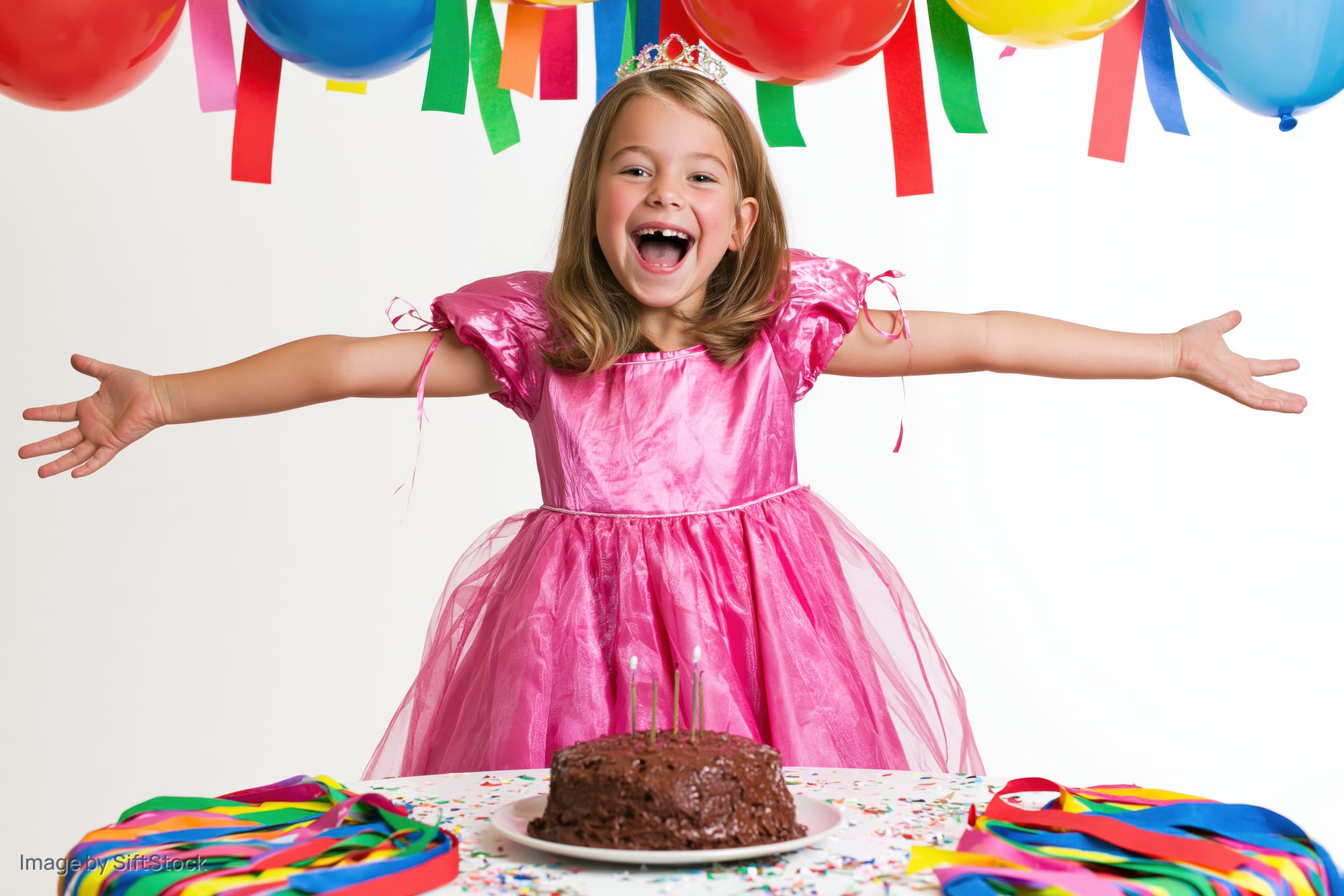 Young girl in pink princess dress smiling with arms open at birthday party table with cake and balloons