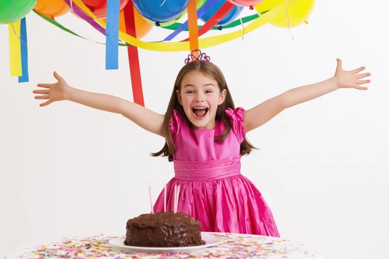 Young girl in pink dress celebrating birthday with arms open, cake and balloons above
