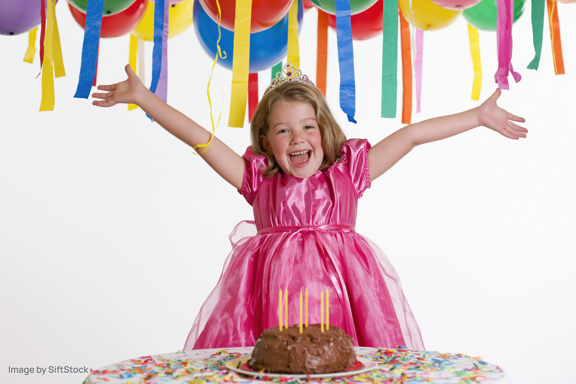 Young girl in pink princess dress with arms raised at birthday party table with cake and balloons