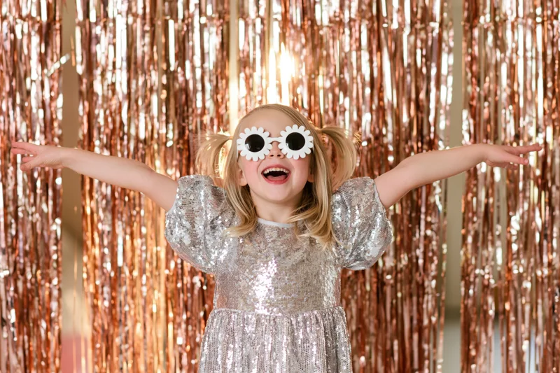 Young girl in silver sequin dress with flower sunglasses smiling with arms open in front of rose gold tinsel curtain