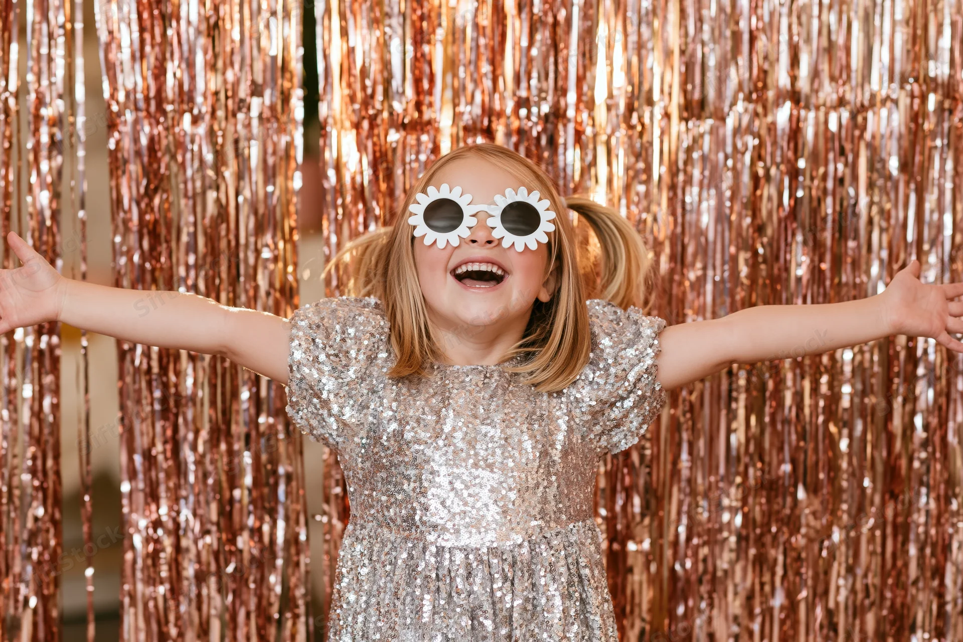Young girl smiling with arms open in front of a gold tinsel curtain wearing a silver sequin dress and white flower sunglasses