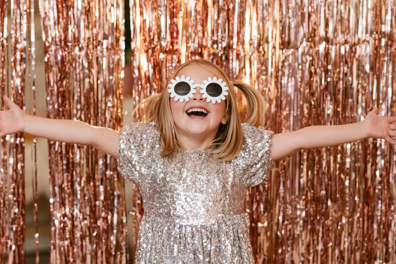Young girl smiling with arms open in front of a gold tinsel curtain wearing a silver sequin dress and white flower sunglasses