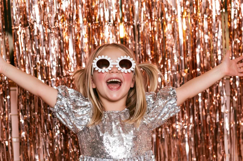 Young girl smiling with arms open in front of copper tinsel backdrop wearing silver sequin dress and flower sunglasses