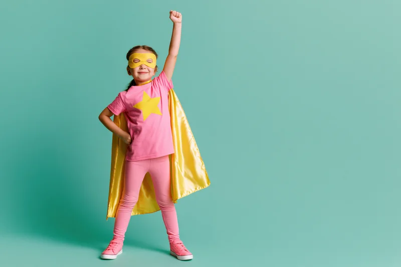 Young girl in pink and yellow superhero costume posing with one fist raised against teal background